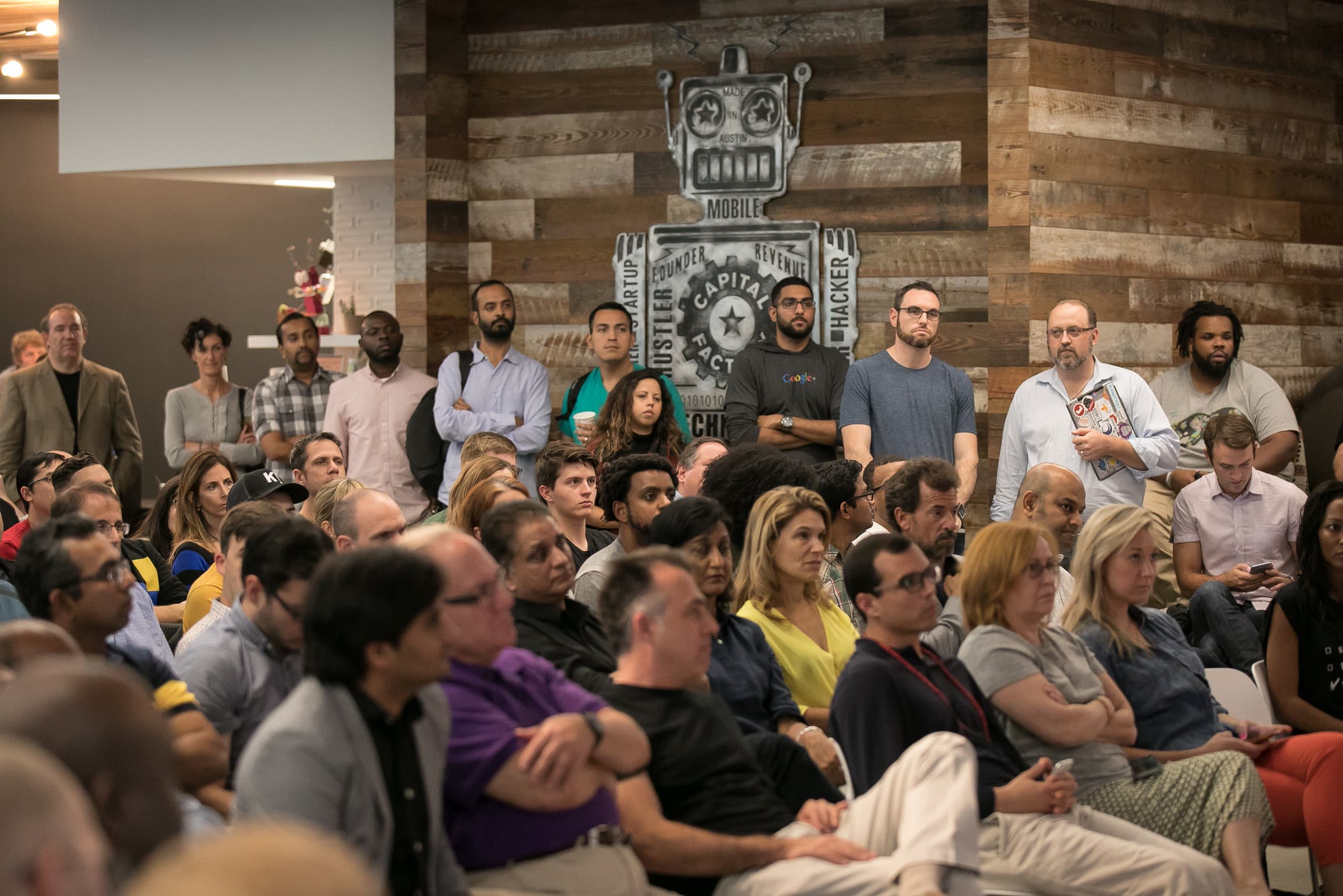 Standing-room demo-day crowd at the reclaimed-wood robot wall